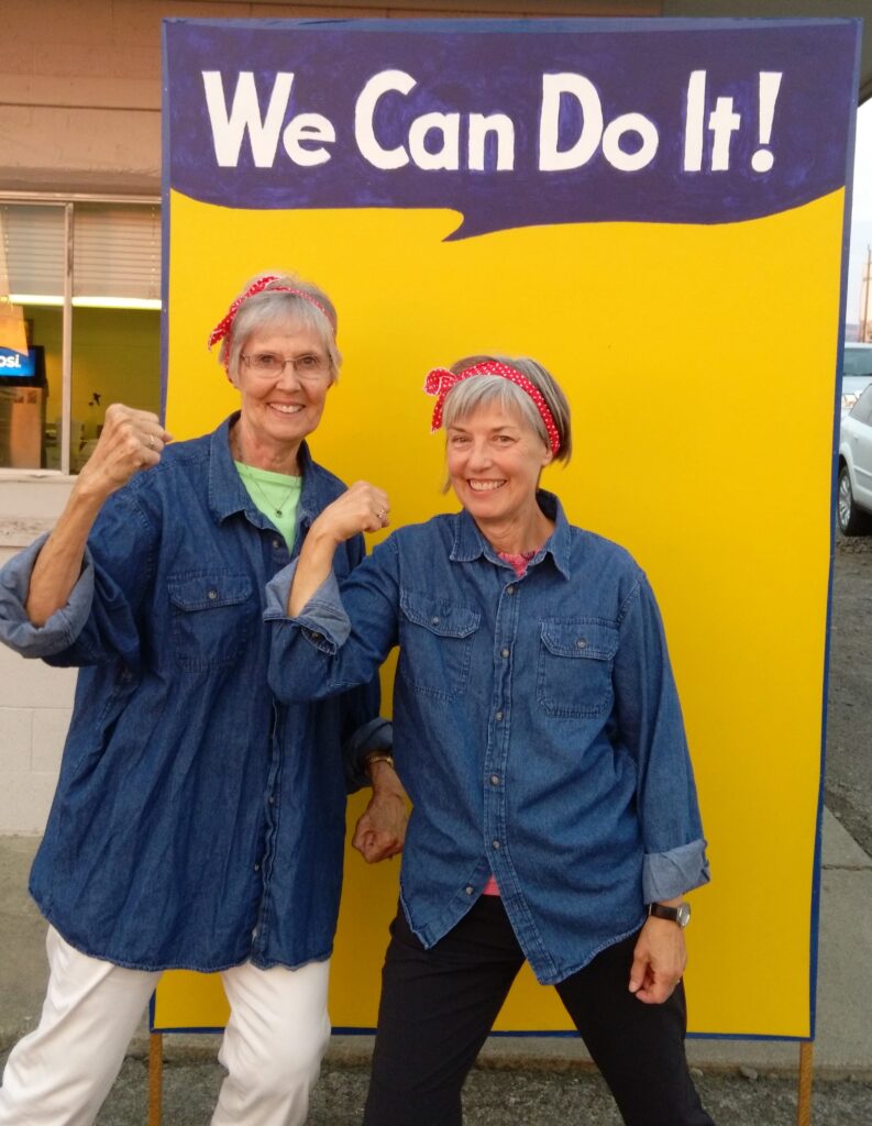 Mary Lynne and Rhonda strike a Rosie the Riveter pose in front of a yellow sign with "We Can Do It!" above their heads.