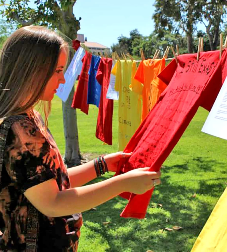 Girl standing by clothesline of multi-colored t-shirts examines a red one with slightly visible writing on it.